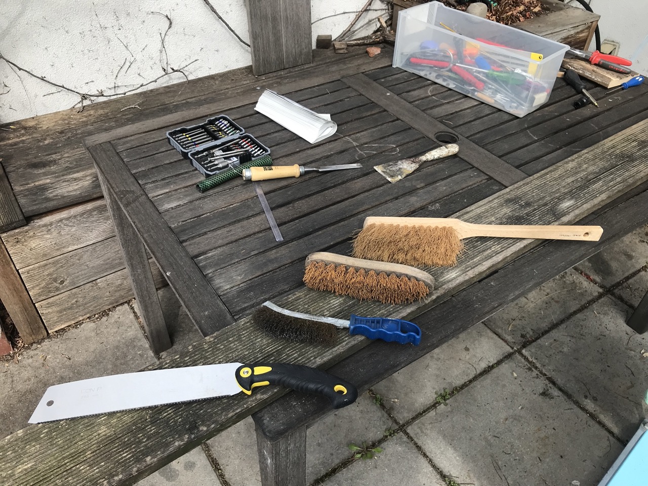 Auf einem verwitterterten Brett liegen eine Japansäge, eine Drahtbürste, eine Wurzelbürste und ein Handbesen. Im Hintergrund diverses anderes Werkzeug.