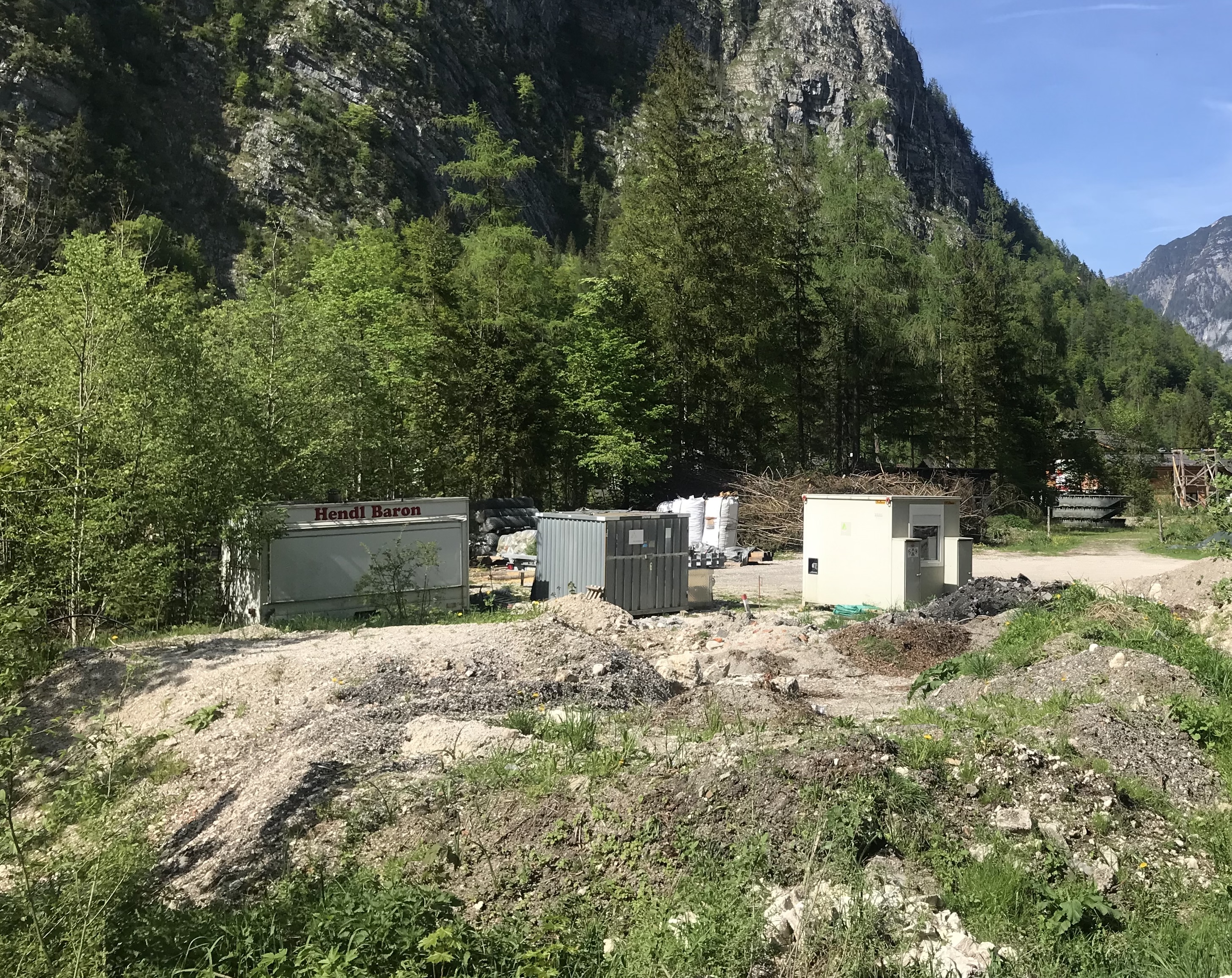 Vor einer Wald- und Bergkulisse befindet sich eine Art Lagerplatz mit diversen Containern auf einem Schutthaufen. Links im Bild eine offensichtlich ausrangierte Imbissbude mit der Aufschrift „Hendl Baron“.