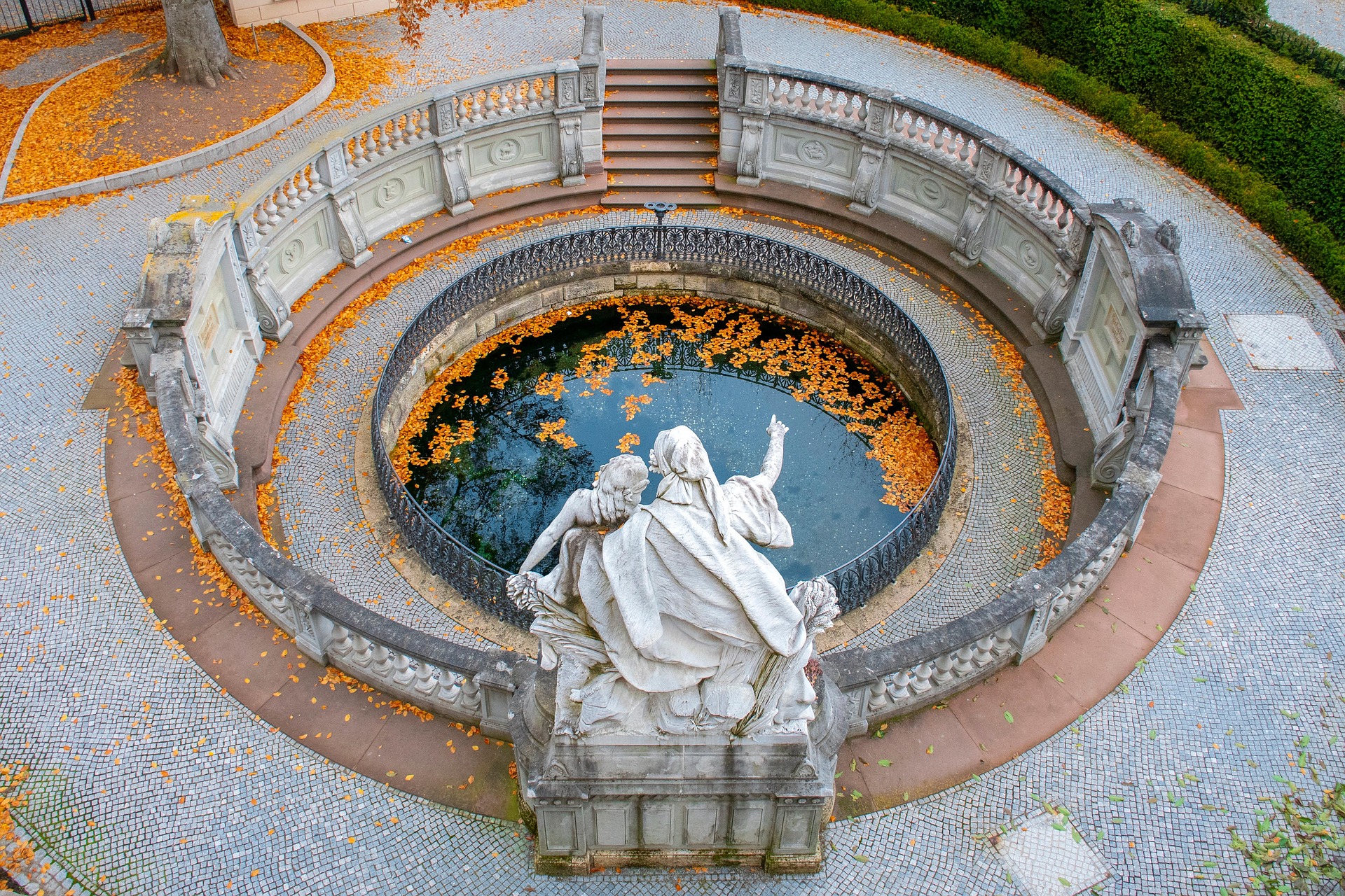 Farbfoto: Donauquelle in Donaueschingen von oben, ein rundes Brunnenbecken mit Herbstlaub auf dem Wasser, umgeben von einer Balustrade, davor eine Figurengruppe aus Marmor